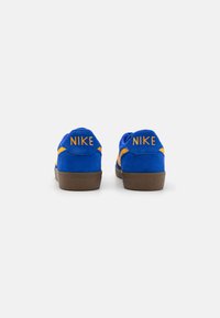 Sneakers azuis em camurça com uma sola de borracha castanha, apresentando detalhes em amarelo e um logotipo na parte de trás. Design de perfil baixo com textura suave.