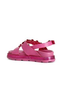 Vaaleanpunaiset korkeat sandaalit ristikkäisillä remmeillä, valmistettu kiiltävästä synteettisestä materiaalista, jossa on tekstuuripohja ja metalliset yksityiskohdat pohjassa.