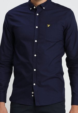 Homme portant une chemise bleu marine ajustée à boutons blancs avec un petit logo jaune brodé sur la poche poitrine.