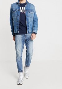 Světle modrá džínová bunda s předními kapsami, oblečená přes námořnický svetr, spárovaná se světle modrými džíny a bílými teniskami se šedými akcenty.