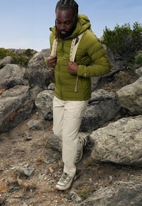 Giacca a vento verde con cappuccio, pantaloni cargo bianchi e scarpe da trekking crema con accenti viola. Sullo sfondo si vedono terreni rocciosi.