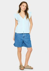 Blusa senza maniche a righe blu con maniche a volant, bottoni frontali, abbinata a pantaloni shorts in denim e sandali beige.
