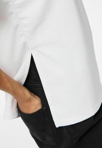 Hvid t-shirt med sideskår, lavet af blød bomuld, præsenteret med en afslappet pasform, båret over sorte denim med en synlig forlommes.