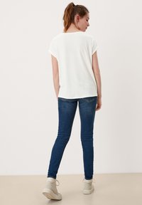 Witte top met korte mouwen, losse pasvorm, gecombineerd met donkerblauwe skinny jeans en lichtbeige sneakers, gezien vanuit de achterkant. Glad stoftextuur.