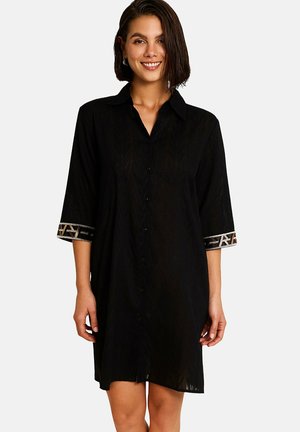 Femme portant une robe chemisier noire à manches trois-quarts avec des poignets à motif géométrique, souriante sur fond blanc.