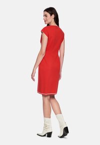 Rotes figurbetontes Kleid mit Kurzärmeln, weißem Besatz am Ausschnitt und Saum, kombiniert mit weißen Stiefeletten mit Absatz. Glatte Textur und schlichtes Design.