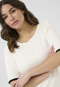 Haut en tricot côtelé couleur crème avec des manches courtes ; présente des bordures noires aux poignets et un col arrondi. Texture lisse, design ajusté.