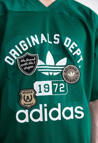 Zöld hálós mez, fehér nyomtatott szöveggel, Adidas logóval és különféle foltokkal, amelyek szöveget és emblémákat tartalmaznak. Néhány folton repedezett textúra található.