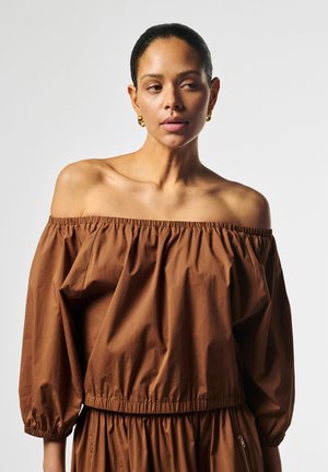 Frau mit zurückgebundenen dunklen Haaren, die goldene Creolen trägt und eine braune schulterfreie Bluse mit langen Ärmeln anhat, schaut zur Seite.