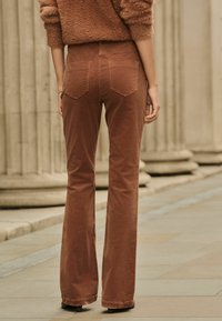 Pantalons en velours côtelé marron évasés avec un design subtil de cinq poches et des coutures marquées, accompagnés d'un pull marron texturé.