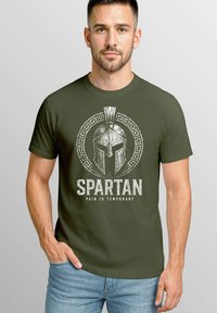 Olive green cotton t-shirt featuring a distressed Spartan helmet graphic and text "SPARTAN PAIN IS TEMPORARY" in white. Short sleeves, crew neck.