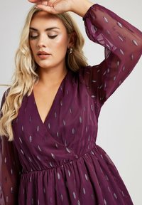 Abito bordeaux con scollatura a V, maniche lunghe trasparenti, vita elastica e motivi argentati simili a piume. Tessuto morbido e leggero.