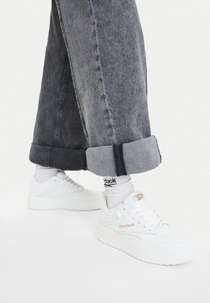 Białe sneakersy Reebok z fakturowanej skóry, sznurowane, z złotym logo. Połączone z szarymi jeansami z podwiniętymi nogawkami i białymi skarpetkami.