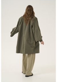 Olive roheline ülev coat, millel on laiad varrukad, lahtine lõige ja nupud mansettides, koos heledate beežide laiad püksid.