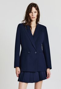 Blazer doppiopetto blu navy con revers a punte, due bottoni e taschino sul petto. Indossato con una minigonna plissettata blu navy.
