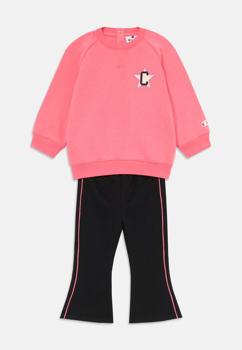 Rozā garām piedurknēm svītra ar zvaigznīti un "C" aplikāciju, pārī ar melnām platām biksēm, kurām ir rozā sānu svītras, novietota uz baltas fona.