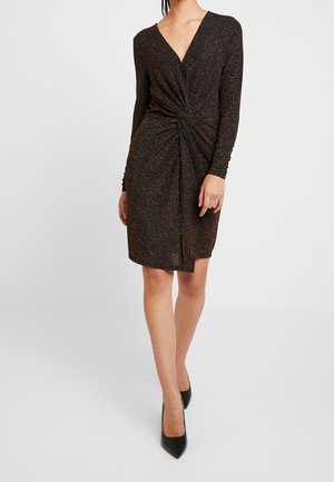 Frau trägt ein schwarzes Kleid mit langen Ärmeln und Glitzerdetails, mit einer gedrehten Raffung an der Taille und schwarzen spitzen Schuhen, steht auf weißem Boden.
