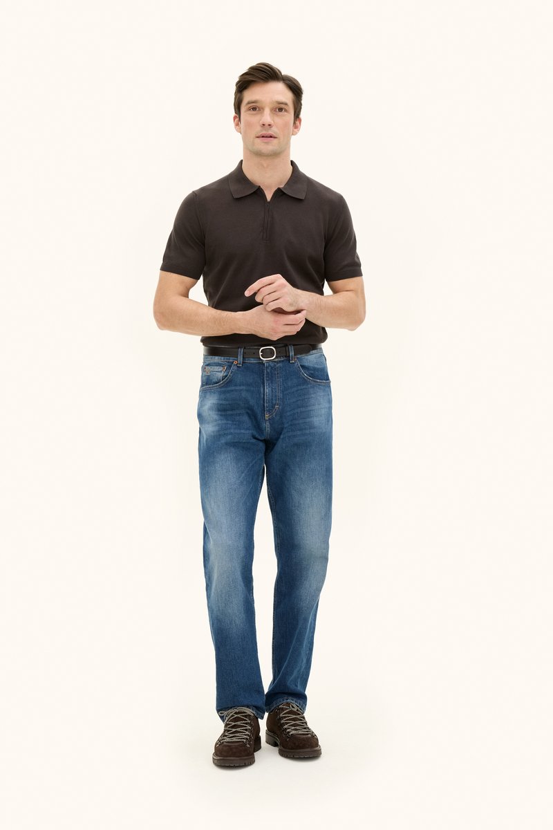 Pikétröja i mörkbrunt, blå jeans i denim och bruna vandringsskor med snören. Modellen står upprätt med händerna tillsammans vid midjan.