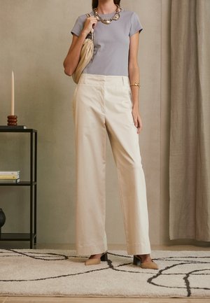 Femme portant un pantalon large beige, un top à manches courtes violet clair, un collier en chaîne doré et un bracelet, tenant un sac à main beige à l'intérieur.