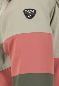 Chaqueta con bloques de color que presenta paneles en verde oliva, rosa coral y topo. Incluye un parche del logo ZigZag triangular en negro. Texto de tejido suave.