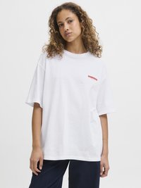 White oversized t-shirt, cotton fabric, short sleeves, crew neck, features red "SOURDOUGH" text on the left side, paired with dark denim pants.