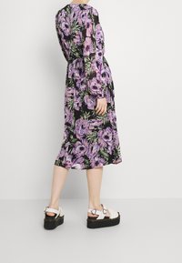 Květinové midi šaty s černým pozadím, zdobené fialovými a zelenými květinami. Dlouhé rukávy, elastický pas, doplněné bílými platformovými sandály.