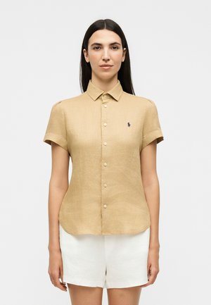 Γυναίκα με μακριά σκούρα μαλλιά, φορώντας μια μπεζ κοντομάνικη πουκαμισά και λευκά σορτς, στέκεται μπροστά σε απλό φωτεινό φόντο.