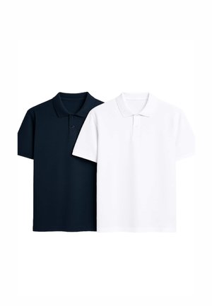 Młody mężczyzna uśmiechający się, ubrany w granatową kurtkę na białej koszulce polo, jasnoniebieskie dżinsy z podwiniętymi mankietami oraz białe tenisówki z czarnymi akcentami.