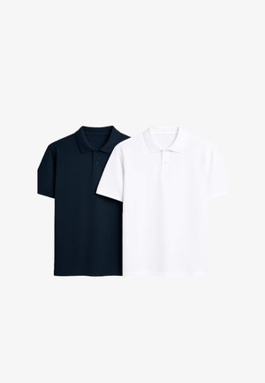 Giovane uomo sorridente, con una giacca blu navy sopra una polo bianca, jeans azzurri con risvolti e sneakers bianche con dettagli neri.