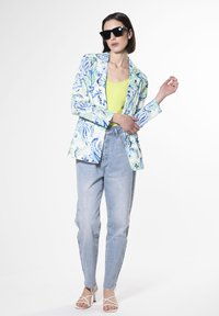Blazer stampato in blu chiaro, verde e bianco, abbinato a una maglietta gialla neon, jeans blu chiaro e tacchi bianchi con cinturini.