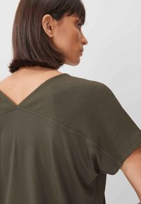 Top verde oliva con schiena a V, maniche corte e texture liscia. Presenta un design pulito e cuciture a contrasto lungo i bordi.