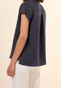 Ciemnoszara lniana koszula z krótkim rękawem, kołnierzykiem i widocznym szwem. Charakteryzuje się luźnym krojem i bocznymi rozcięciami dla swobody ruchów.