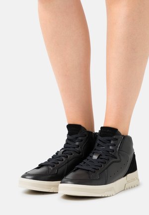 Baskets montantes noires avec semelles blanches portées sur des jambes nues sur fond blanc, montrant les vues latérale et avant.