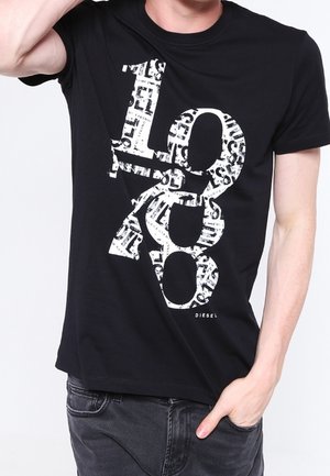 Personne portant un t-shirt Diesel noir avec les grands chiffres blancs 1978 imprimés à l'avant, main dans la poche.