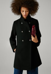Melna vilnas mēteli ar divrindu dizainu, sudraba pogām un sānu kabatām. Modelis tur sarkanu rokassomu.