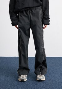 Černé sportovní kalhoty s volným střihem, elastickým pasem a kontrastním bílým proužkem po stranách. Kombinovány se šedými teniskami.