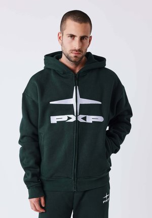 Jeune homme aux cheveux courts portant un sweat à capuche zippé vert foncé et un pantalon avec le logo blanc PXP, debout devant un fond uni.