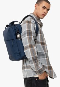 Marineblauer Rucksack mit gepolsterten Trägern und einem Frontklappenverschluss, der eine silberne Wasserflasche hält, getragen über einem karierten grauen und braunen Hemd.