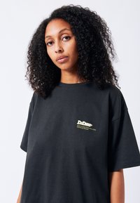 Černé bavlněné tričko s krátkým rukávem, volný střih, s malým žlutým logem na levém prsou. Hladká textura a jednoduchý design.