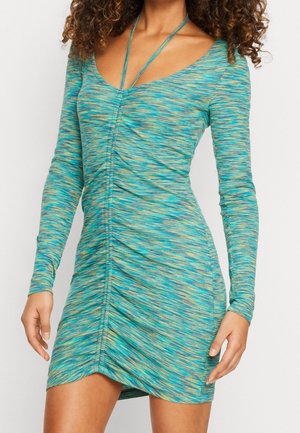 Vrouw draagt een nauwsluitende jurk met lange mouwen, geplooid, met een blauwgroen abstract patroon en een diepe V-hals met een halterbandje.