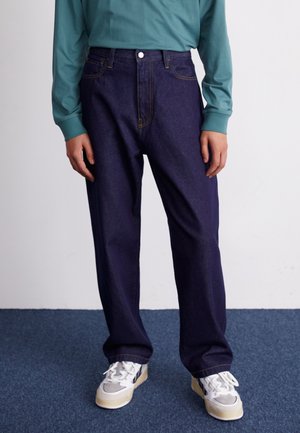 Personne portant un jean bleu foncé ample, une chemise à manches longues bleu sarcelle et des baskets blanches, debout sur un tapis bleu contre un mur blanc.