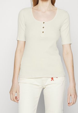 Crème kleurige ribgebreide top met korte mouwen en een ronde halslijn, versierd met drie gouden knopen. Gecombineerd met lichte broeken met rode borduurdetails.