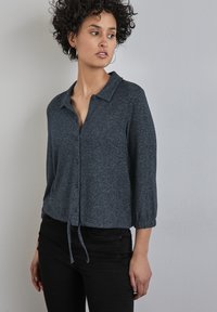 Mörkgrå knäppt blus med krage, trekvartsärmar och dragsko i nederkant. Mjuk, strukturerad tyg med en melerad mönstring.