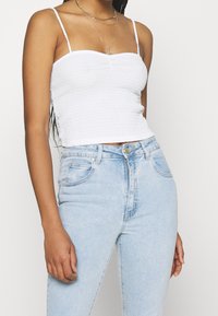 Top débardeur ajusté en maille blanche avec des bretelles fines, associé à un jean taille haute bleu clair avec une fermeture éclair et un bouton.