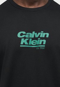 Κοντινό πλάνο μαύρου πουκάμισου με τιρκουάζ κείμενο "Calvin Klein" και την επιγραφή "Est. 1968" κάτω από το στήθος.