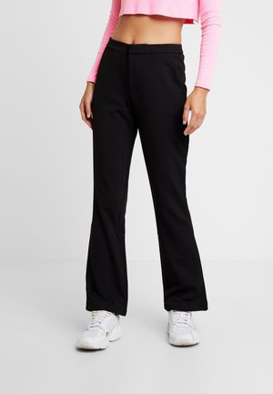 Persoon met zwarte broek met rechte pijpen, witte sneakers en een korte, lange roze top, staand tegen een witte achtergrond.