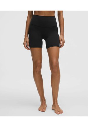 Femme portant un short noir taille haute moulant, debout pieds nus sur un fond uni gris clair.