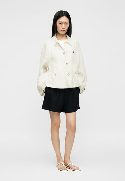 Žena s rovnými čiernymi vlasmi, ktorá má na sebe bielu zapínanú bundu, čierne šortky a biele žabky, stojaca s rukami v kapsách na jednofarebnom pozadí.