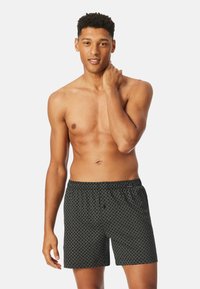 Black boxer shorts with a white geometric pattern, elastic waistband, and relaxed fit. Model has a medium build and is shirtless.