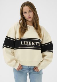 Sweat-shirt couleur crème avec des rayures noires et le texte "LIBERTY UNIVERSITY". Présente des épaules tombantes et des poignets côtelés pour une texture.